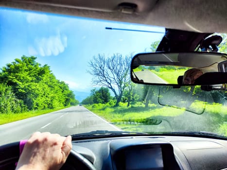 Car salon, windshield, hand of woman on steering wheel and landscape. View from seat of driver on nature with Road, tree and blue sky at sunny day