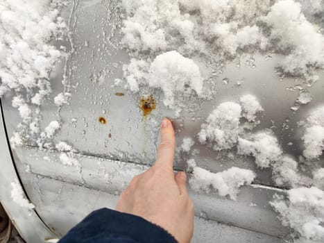 A person points at visible rust spots on a car's exterior covered with snow during cold weather in a neighborhood.
