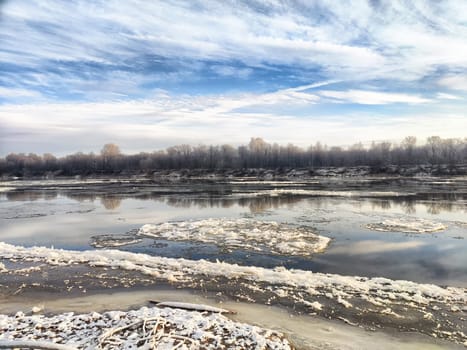 The river showcases large ice floes drifting gently as spring emerges, revealing clear water and a changing landscape.