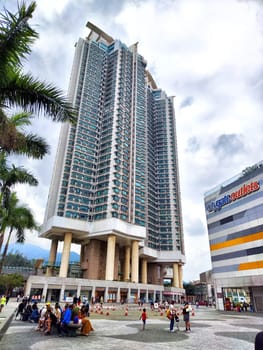 Hong Kong, China - April 08, 2024 - Busy crowds gather at the base of a striking skyscraper as modern architecture rises against the cloudy sky in Hong Kong.