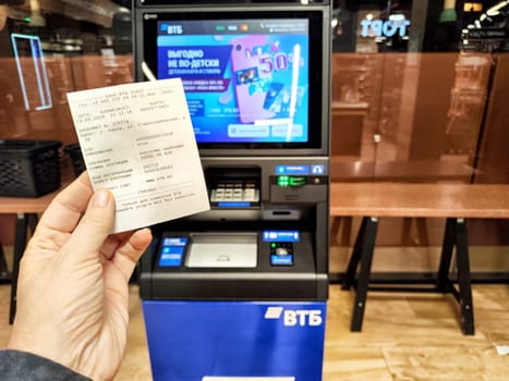 Kirov, Russia - September 29, 2024: Individual holds a receipt after completing a cash withdrawal at an automated teller machine in a busy interior setting.