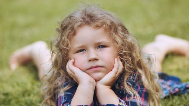 Curly haired blond girl resting on green grass, hands covering face, relaxing during family outdoor moment