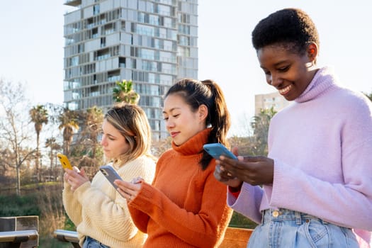 Three young women are outdoors, joyfully engaging with their smartphones while soaking in a vibrant urban setting