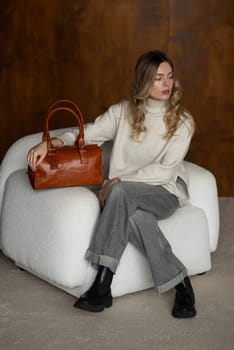 beautiful girl in gray pants and beige sweater posing with a small brown leather bag.
