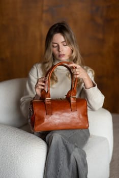 beautiful girl in gray pants and beige sweater posing with a small brown leather bag.