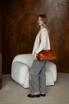 beautiful girl in gray pants and beige sweater posing with a small brown leather bag.