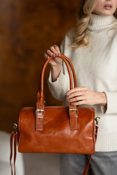 beautiful girl in gray pants and beige sweater posing with a small brown leather bag.