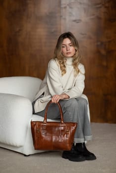 beautiful girl in gray pants and beige sweater posing with a small brown leather bag.