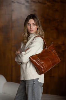 beautiful girl in gray pants and beige sweater posing with a small brown leather bag.