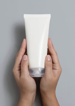 Female hands holding white squeeze cream tube against grey top view, cosmetic package mockup, copy space. Skincare beauty product