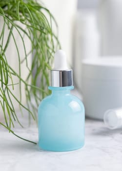 Light blue glass dropper bottle on white marble near potted green plant and cosmetics in bathroom, close up, packaging mockup. Natural product for facecare, serum or essence
