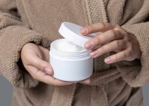 Female hands with natural manicure opening white blank cream jar, cosmetic mockup. Close up of woman in bathroom wearing light brown bathrobe. Bodycare concept