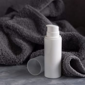 White blank one pump cream bottle near dark grey towel in bathroom closeup, brand packaging mockup, copy space. Natural handmade cosmetic product for facecare and spa procedures