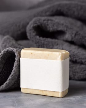 Beige handmade soap bar with blank label near dark grey folded towel closeup, packaging mockup, copy space. Natural herbal hygiene soap for daily cleansing and skincare