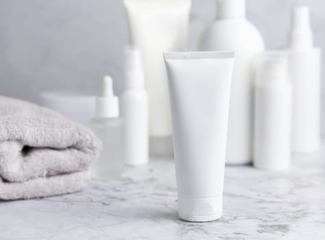 White cream squeeze tube on white marble table against cosmetic bottles and grey towel in bathroom, close up, packaging mockup. Natural product for daily face care, spa procedures