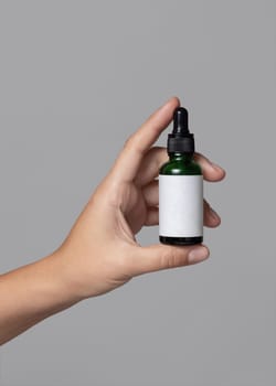 Female hand holding green glass dropper bottle with blank label against grey close up, cosmetic packaging mockup, copy space. Skincare beauty product - serum or essential oil