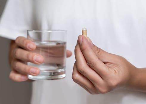 Woman in white t-shirt holding beige capsule in fingers and water glass in hand closeup. Female or male taking dietary supplement, daily vitamins. Healthcare concept
