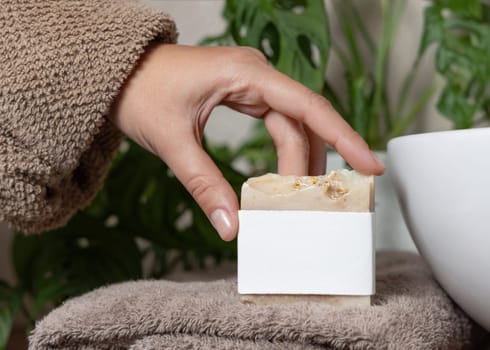 Female hands with natural manicure holding soap bar with blank label, packaging mockup. Close up of woman in bathroom wearing light brown bathrobe agains green plant. Hygiene concept