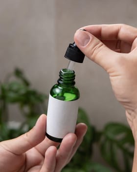 Female hands with natural manicure opening green dropper bottle with blank label, close up against green plant. Mockup for cosmetic bottle with pipette, serum or essence. Beauty spa spa procedures