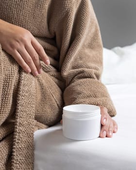 Woman in brown bathrobe puts white opened cream jar on white bedsheet closeup. Bodycare concept. Organic cosmetics for body and face care, beauty and spa procedures