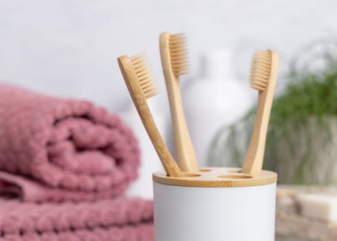 Eco-friendly wooden bamboo toothbrushes in holder cup in a white in bathroom against natural cosmetics, towel and green plant, mockup. Personal care and hygiene products