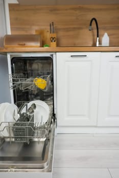 Vertical photography an open dishwasher with a drawer pulled out close-up on the left, it contains clean dishes and cutlery against the rest of the kitchen in a white and wooden style, kitchen utensils and a faucet with sink visible. Comfort concept, dishwashing. High quality photo