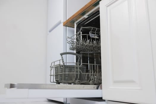 Side view of an empty open dishwasher with gray drawers for dishes and cutlery stands against the background of white kitchen cabinets and a white refrigerator. Dishwashing concept, comfort. High quality photo