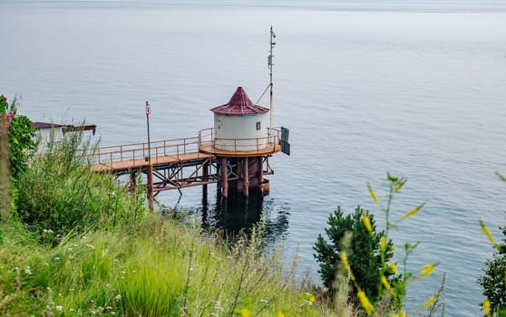 Small white water intake building standing on stilts in a calm russian lake, connected to the shore by a metal pier and surrounded by lush vegetation on an overcast day