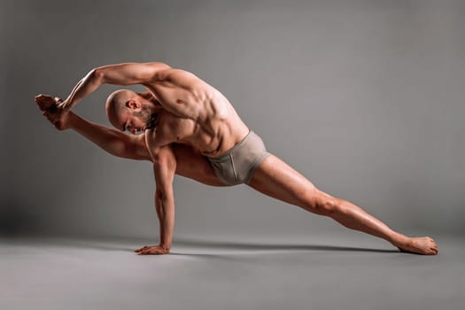 Muscular male dancer is doing the splits while holding his foot during a dance practice over a gray studio background