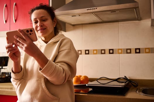A cheerful woman wearing casual attire uses her smartphone while standing in her kitchen. The setting features a modern design with a backsplash and nearby kitchen utensils.