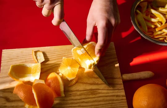 A close-up view of hands slicing a peeled orange on a wooden cutting board, emphasizing fresh fruits and the culinary process against a vivid red background.