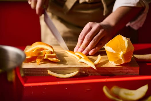 A person carefully slices an orange peel on a wooden cutting board, capturing culinary preparation.