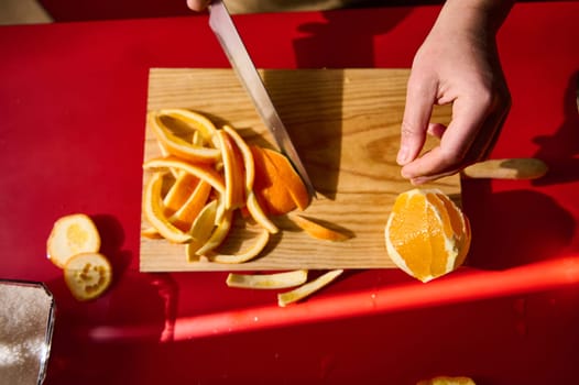 A hand seen peeling and slicing an orange on a cutting board, surrounded by an assortment of orange peels on a vibrant red surface.