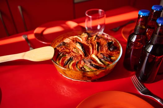 A freshly baked ratatouille served on a vibrant red table alongside rustic drink bottles.