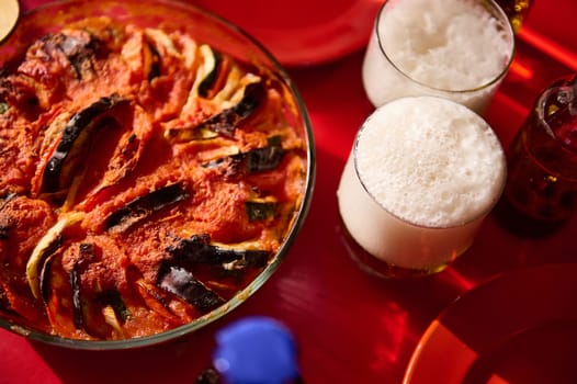 A vibrant baked vegetable casserole served on a red table, accompanied by two foamy beer glasses. The image captures a moment of delicious food and refreshing drinks, emphasizing warmth, enjoyment, and sociability.