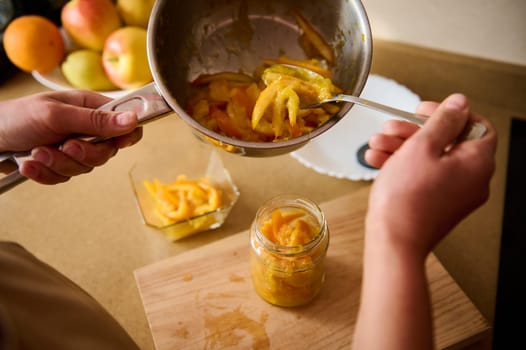 A person transfers freshly cooked, sliced fruits into a jar to make preserves. Fresh fruits and a clean wooden surface accompany the preparation in a cozy kitchen setting.