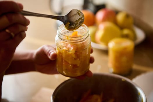 Hands carefully filling glass jars with sliced fruit and spices, preparing homemade preserves in a kitchen. A background of fresh ingredients creates a warm and productive atmosphere.