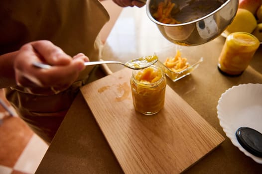 A close-up of a person using a spoon to prepare fruit jam in jars, showcasing a homemade and artisan culinary process in a kitchen environment.