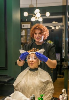 A young girl is getting her hair curled with curlers by a hairdresser. The client is sitting in a chair in a beauty salon. Her hair is being styled for the hair curling procedure