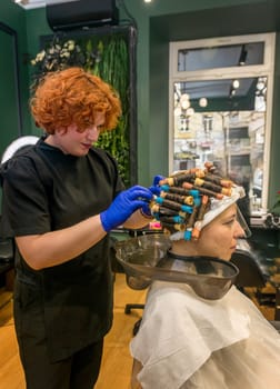 A young girl is getting her hair curled with curlers by a hairdresser. The client is sitting in a chair and her hair is curled with curlers. The hairdresser is curling the curlers