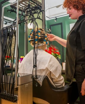 the client is sitting in a chair with curlers on her head the device treats and makes hair curly and wavy. the master adjusts the curlers the device is connected