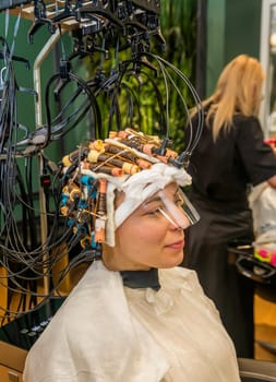 modern process of hair treatment and curling. client sits in chair with curlers on her head. machine treats and makes hair curly and wavy.