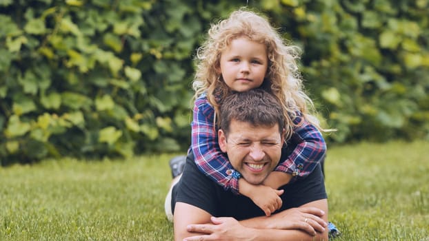 Cheerful girl embracing loving father, resting on grassy ground during warm sunny afternoon, sharing tender family moment