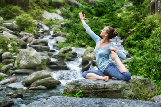 Young sporty fit woman doing yoga asana Eka pada rajakapotasana - one-legged king pigeon pose at tropical waterfall