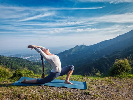 Sporty fit woman practices yoga Anjaneyasana - low crescent lunge pose outdoors in mountains in morning