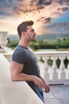 A man standing on a balcony next to a white railing. Photo of a muscular and handsome man standing on a balcony next to a white railing