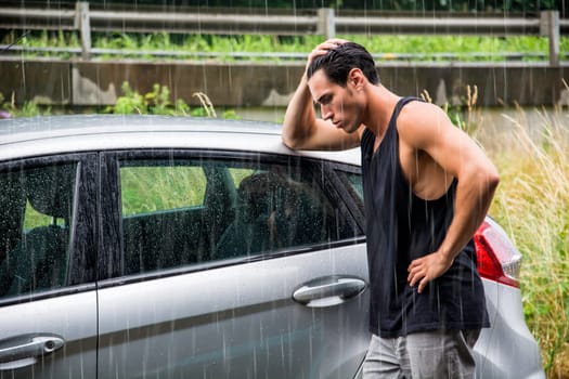 A man standing next to a car in the rain, sad because his vehicle broke down