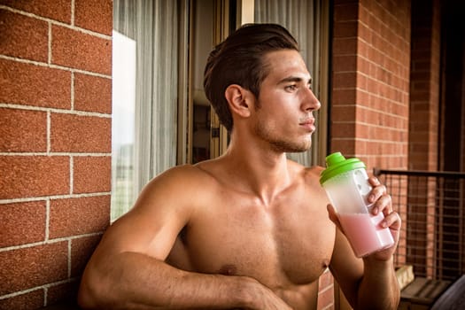 young and attractive muscular shirtless man enjoying a colorful protein shake, sitting outdoor next to a brick wall