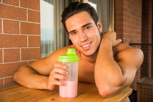 young and attractive muscular shirtless man enjoying a colorful protein shake, sitting outdoor next to a brick wall