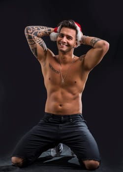 A shirtless man with a santa claus hat on. Photo of a shirtless man wearing a Santa Claus hat in studio, kneeling on the ground on black background